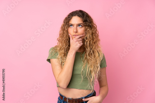 Young blonde woman with curly hair isolated on pink background having doubts and with confuse face expression
