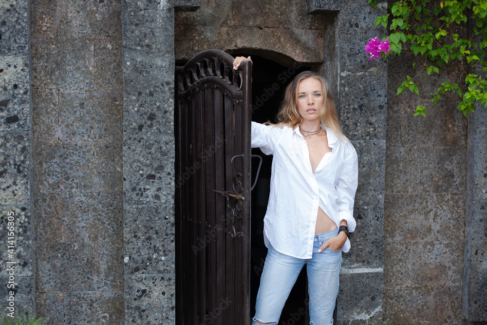 beautiful slender blonde girl with long hair in a white shirt and blue jeans stands at the door of an old stone house