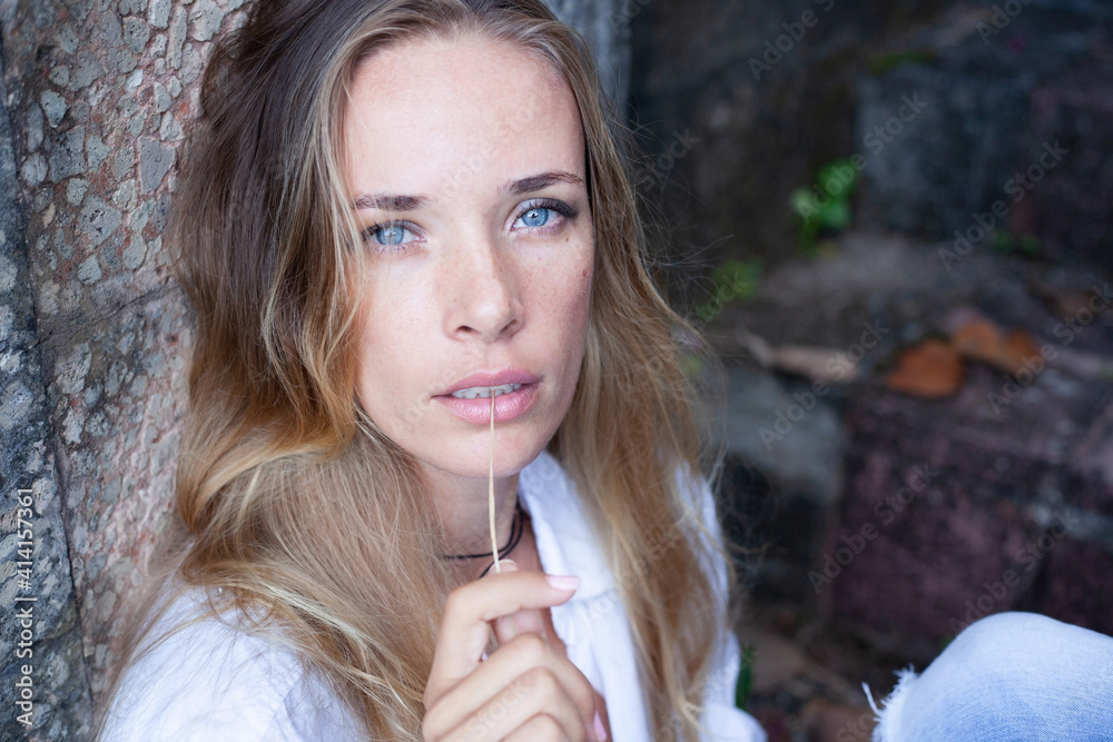 close-up beautiful slender blonde girl with long hair in white shirt sits close to old rastie stone wall and holds straw in mouth close-up beautiful slender blonde girl with long hair in white shirt sits close to old rastie stone wall and holds straw in mouth
