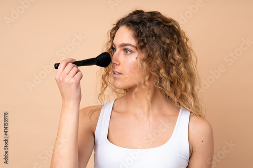 Young blonde woman with curly hair isolated on beige background holding makeup brush