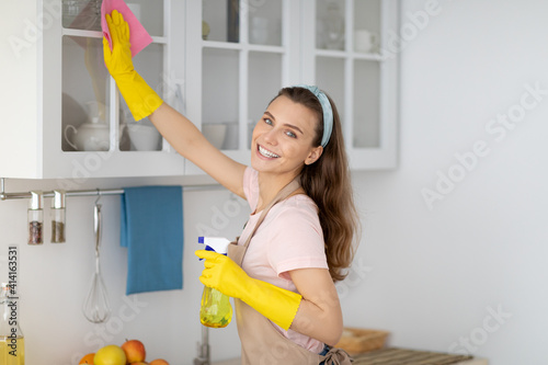 Positive female housekeeper tidying modern apartment, wiping kitchen cabinat with detergent and rag photo