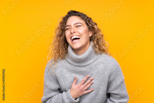 Young blonde woman with curly hair wearing a turtleneck sweater isolated on yellow background smiling a lot © luismolinero