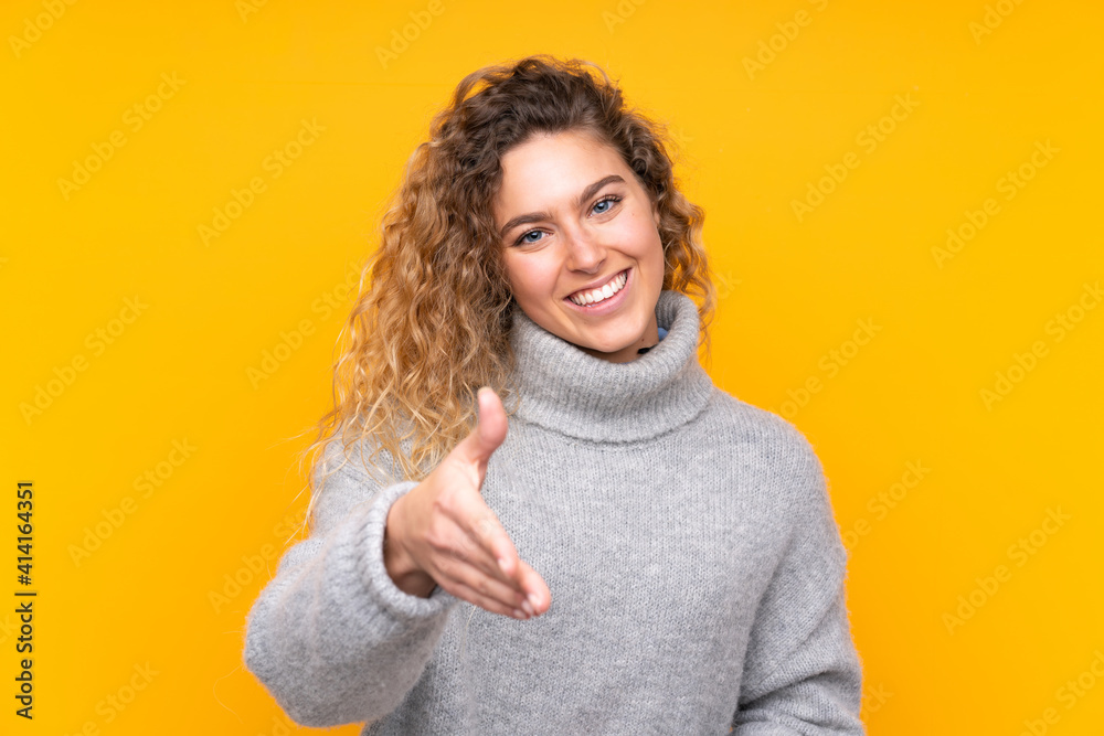Young blonde woman with curly hair wearing a turtleneck sweater isolated on yellow background shaking hands for closing a good deal Young blonde woman with curly hair wearing a turtleneck sweater isolated on yellow background shaking hands for closing a good deal