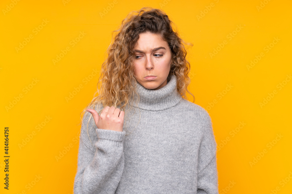 Young blonde woman with curly hair wearing a turtleneck sweater isolated on yellow background unhappy and pointing to the side Young blonde woman with curly hair wearing a turtleneck sweater isolated on yellow background unhappy and pointing to the side