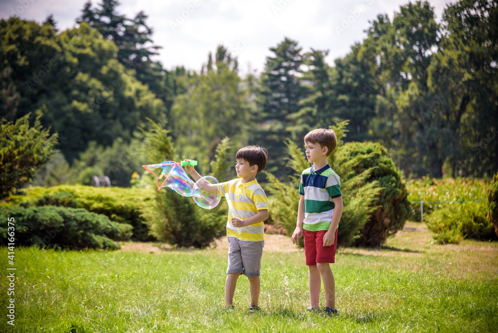 Fototapeta premium Boy blowing soap bubbles while an excited kid enjoys the bubbles. Happy teenage boy and his brother in a park enjoying making soap bubbles. Happy childhood friendship concept
