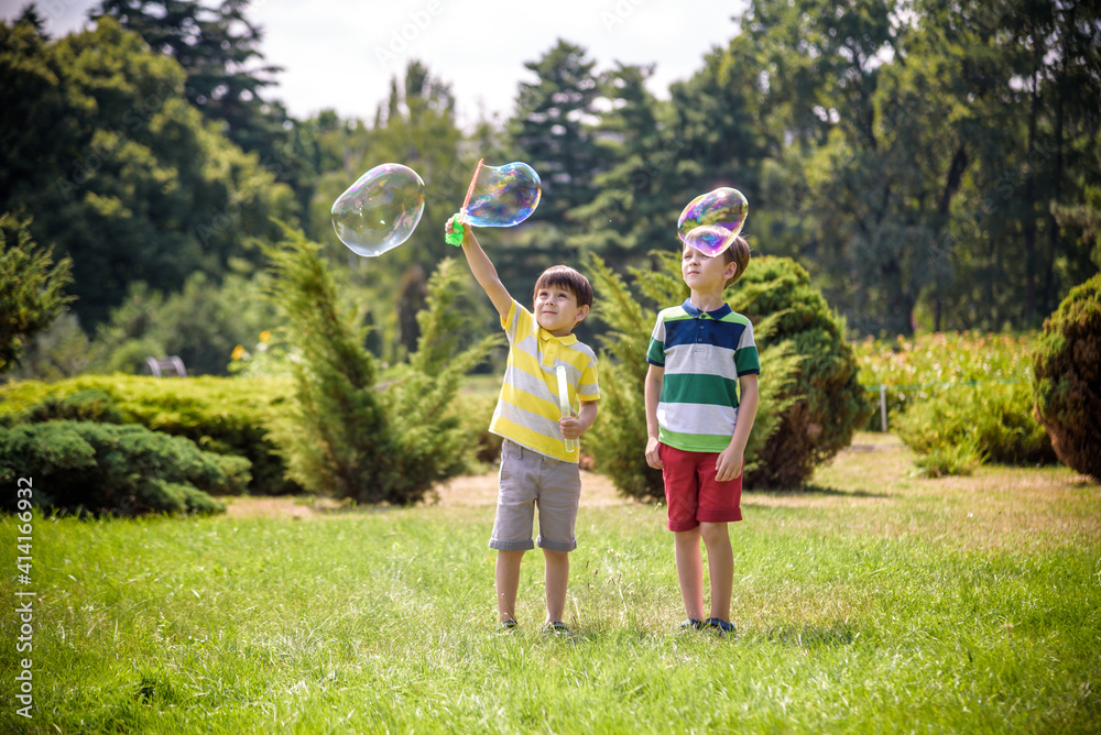 Fototapeta premium Boy blowing soap bubbles while an excited kid enjoys the bubbles. Happy teenage boy and his brother in a park enjoying making soap bubbles. Happy childhood friendship concept