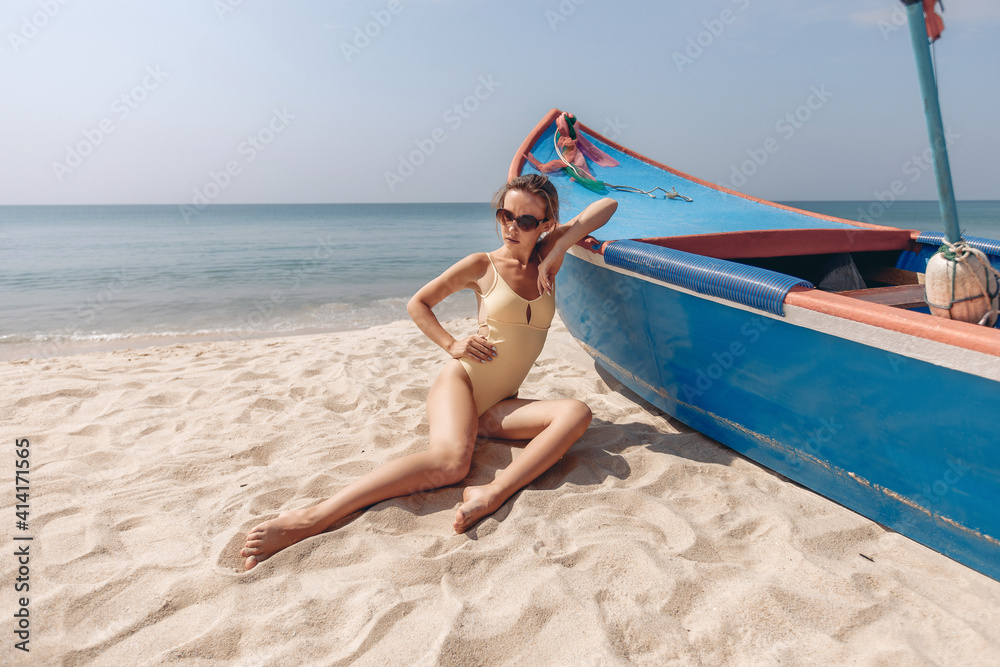 Cute young and independent model with a perfect figure, wearing a bikini, sunglasses and braided hair, lying and posing on the sand of her favorite beach, leaning her elbow on the boat. Cute young and independent model with a perfect figure, wearing a bikini, sunglasses and braided hair, lying and posing on the sand of her favorite beach, leaning her elbow on the boat.
