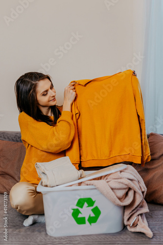 Obraz na płótnie Woman sorting old clothes into white container with recycle sign. Green recycling sign on white container. Woman throwing out old clothes. w salonie