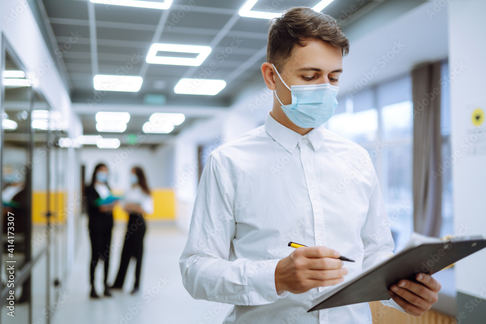 Portrait of young business man in protective face mask an office building hallway. Office worker working in the office during pandemic in quarantine city. COVID - 19. Portrait of young business man in protective face mask an office building hallway. Office worker working in the office during pandemic in quarantine city. COVID - 19.