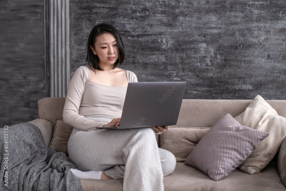 Korean woman holds a laptop in her hands, communicates and works at home on a sofa in a loft interior. Lockdown. Korean woman holds a laptop in her hands, communicates and works at home on a sofa in a loft interior. Lockdown.