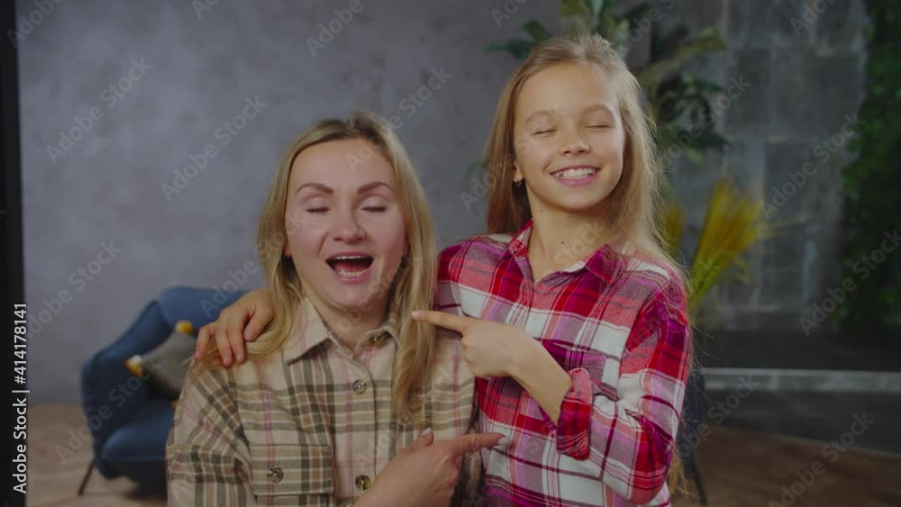 Playful cheerful lovely school age girl and carefree pretty mother with eyes closed playing funny cognitive game, answering for questions and showing with finger who is while enjoying leisure at home.