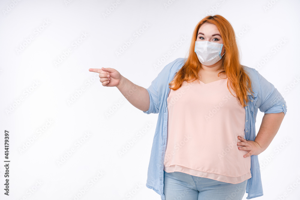 Fat young redhead woman in medical mask against Covid19 pointing at copy space isolated over white background Fat young redhead woman in medical mask against Covid19 pointing at copy space isolated over white background