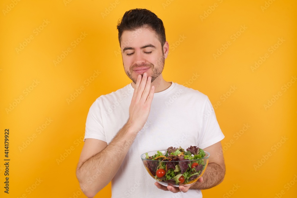 Indoor closeup of young handsome Caucasian man holding a salad bowl against yellow wall practicing yoga and meditation, holding palms together in namaste, looking calm, relaxed and peaceful. Indoor closeup of young handsome Caucasian man holding a salad bowl against yellow wall practicing yoga and meditation, holding palms together in namaste, looking calm, relaxed and peaceful.