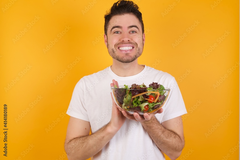 young handsome Caucasian man holding a salad bowl against yellow wall keeps palms together, has pleased expression. Glad attractive male makes request, pleads for mercy. Hopeful young adult. young handsome Caucasian man holding a salad bowl against yellow wall keeps palms together, has pleased expression. Glad attractive male makes request, pleads for mercy. Hopeful young adult.