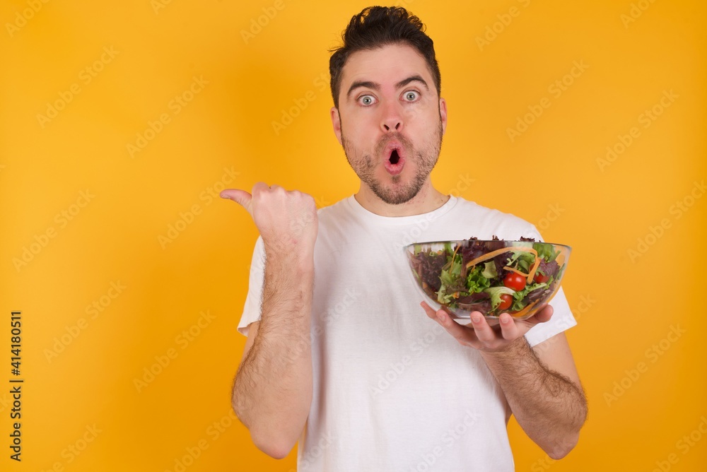 Shocked young handsome Caucasian man holding a salad bowl against yellow wall points with thumb away, indicates something. Check this out. Advertisement concept. Shocked young handsome Caucasian man holding a salad bowl against yellow wall points with thumb away, indicates something. Check this out. Advertisement concept.