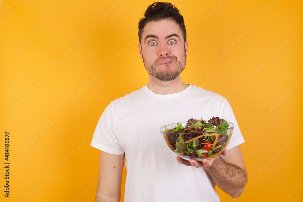 young handsome Caucasian man holding a salad bowl against yellow wall being nervous and scared biting lips looking camera with impatient expression, pensive. young handsome Caucasian man holding a salad bowl against yellow wall being nervous and scared biting lips looking camera with impatient expression, pensive.