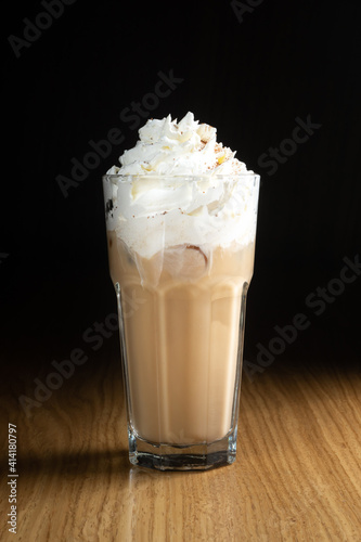 Iced coffee with ice, milk and whipped cream in a tall transparent glass on a wooden table