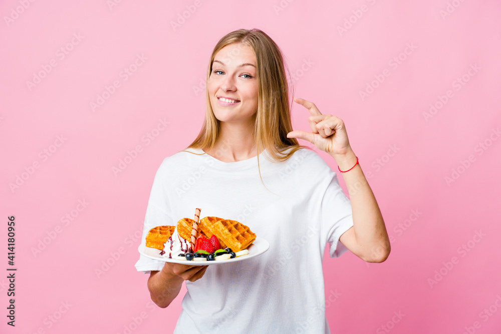 Young russian woman eating a waffle isolated holding something little with forefingers, smiling and confident. Young russian woman eating a waffle isolated holding something little with forefingers, smiling and confident.