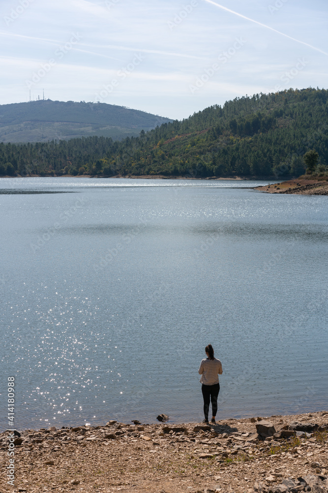 Fototapeta premium Caucasian girl looking at the lake water of Apartadura dam with mountains with trees landscape in Alentejo, Portugal
