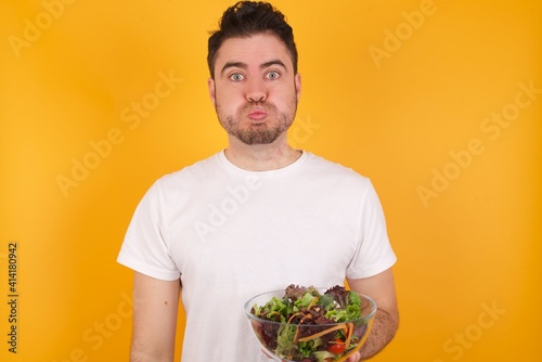 young handsome Caucasian man holding a salad bowl against yellow wall puffing cheeks with funny face. Mouth inflated with air, crazy expression.