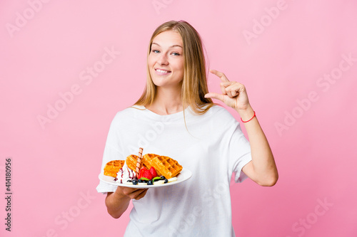 Young russian woman eating a waffle isolated holding something little with forefingers, smiling and confident.