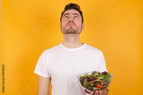 young handsome Caucasian man holding a salad bowl against yellow wall looking up as he sees something strange.