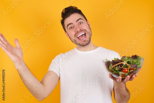 Cheerful young handsome Caucasian man holding a salad bowl against yellow wall making a welcome gesture raising arms over head.