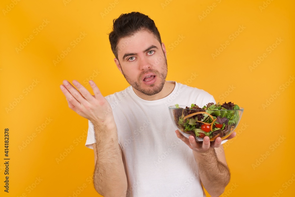 Studio shot of frustrated young handsome Caucasian man holding a salad bowl against yellow wall gesturing with raised palm, frowning, being displeased and confused with dumb question. Studio shot of frustrated young handsome Caucasian man holding a salad bowl against yellow wall gesturing with raised palm, frowning, being displeased and confused with dumb question.