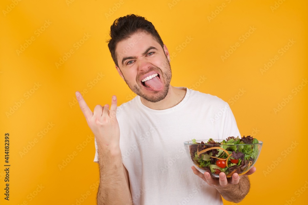 Portrait of a crazy young handsome Caucasian man holding a salad bowl against yellow wall showing tongue horns up gesture, expressing excitement of being on concert of band. Portrait of a crazy young handsome Caucasian man holding a salad bowl against yellow wall showing tongue horns up gesture, expressing excitement of being on concert of band.
