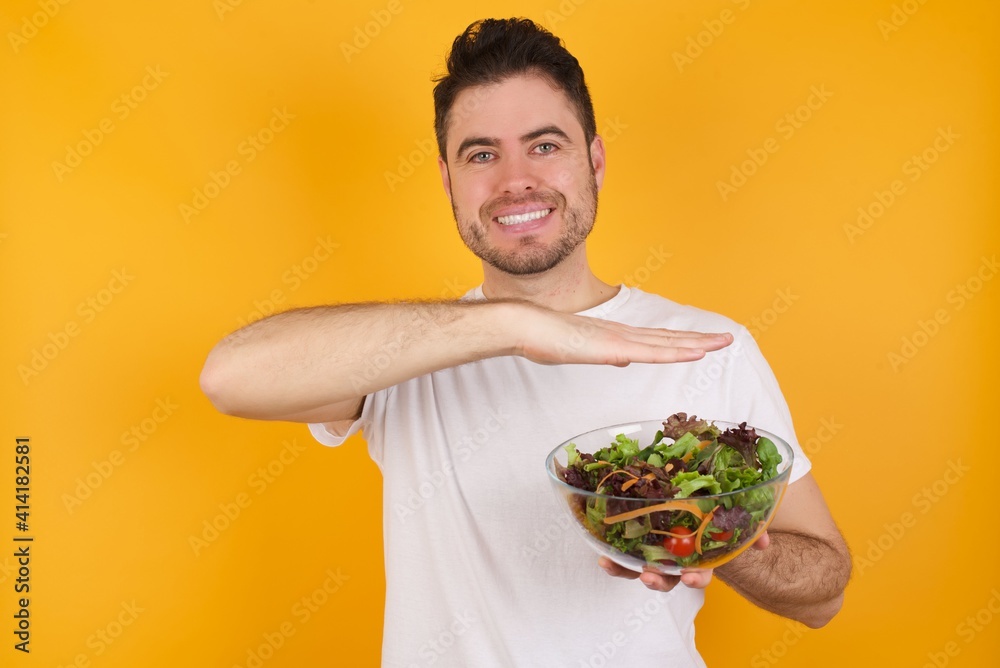 young handsome Caucasian man holding a salad bowl against yellow wall gesturing with hands showing big and large size sign, measure symbol. young handsome Caucasian man holding a salad bowl against yellow wall gesturing with hands showing big and large size sign, measure symbol.