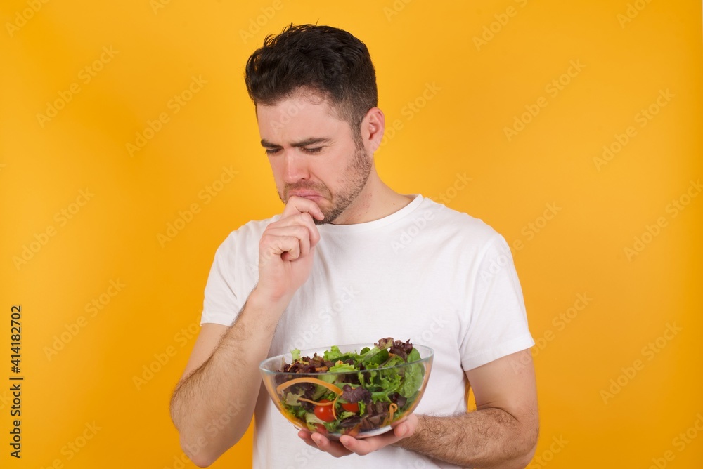 Sad young handsome Caucasian man holding a salad bowl against yellow wall feeling upset while spending time at home alone staring at camera with unhappy or regretful look. Sad young handsome Caucasian man holding a salad bowl against yellow wall feeling upset while spending time at home alone staring at camera with unhappy or regretful look.