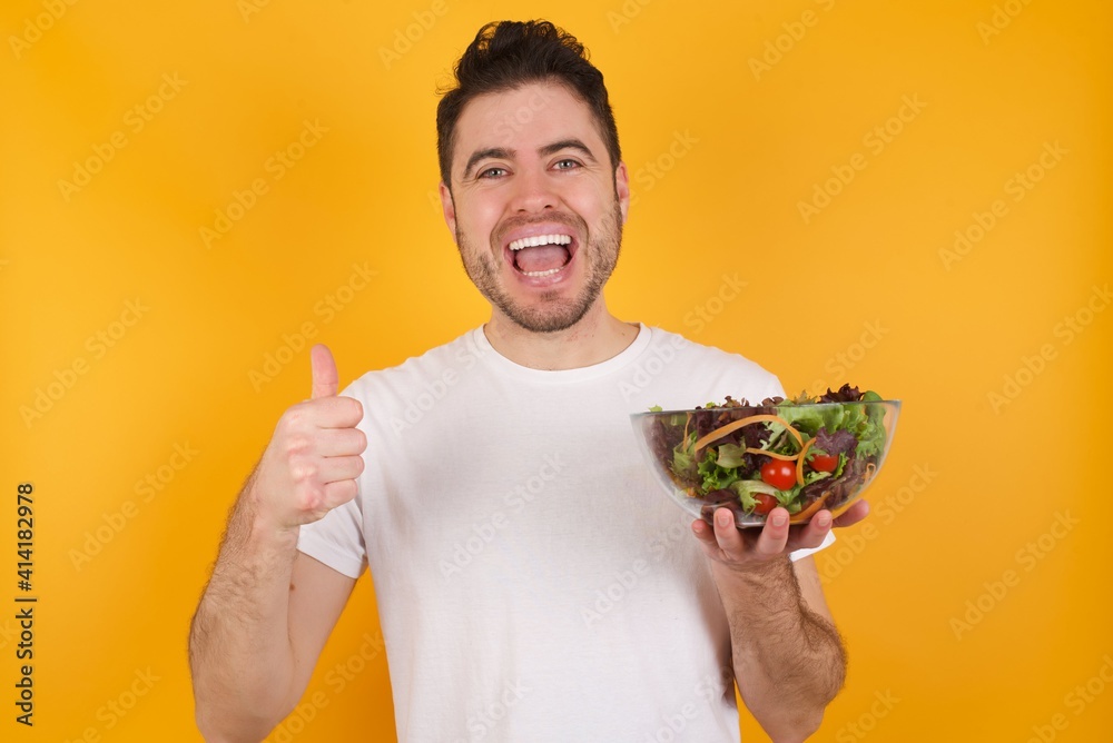 Optimistic young handsome Caucasian man holding a salad bowl against yellow wall showing thumbs up with positive emotions. Quality and recommendation concept. Optimistic young handsome Caucasian man holding a salad bowl against yellow wall showing thumbs up with positive emotions. Quality and recommendation concept.