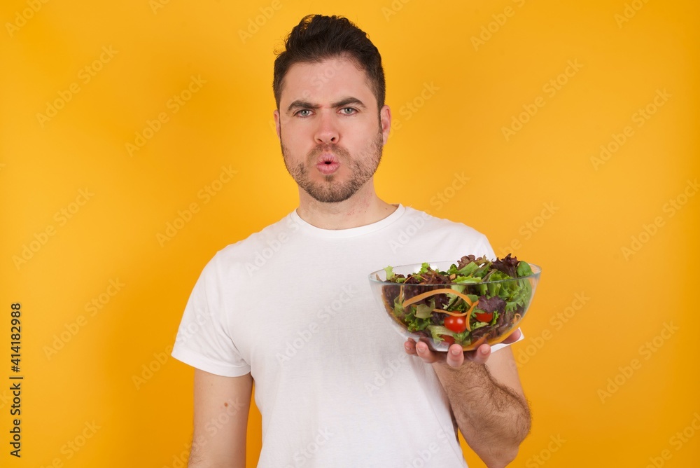 young handsome Caucasian man holding a salad bowl against yellow wall expressing disgust, unwillingness, disregard having tensive look frowning face, looking indignant with something. young handsome Caucasian man holding a salad bowl against yellow wall expressing disgust, unwillingness, disregard having tensive look frowning face, looking indignant with something.