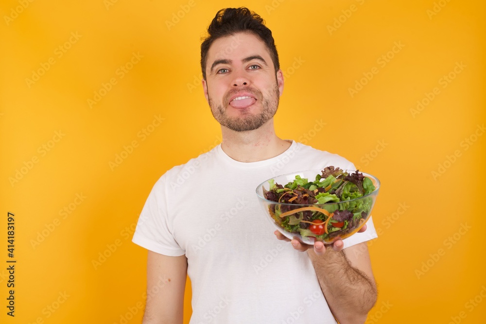 young handsome Caucasian man holding a salad bowl against yellow wall with happy and funny face smiling and showing tongue.