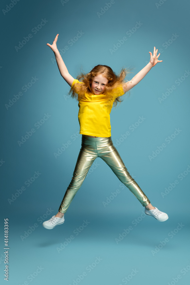 Jumping high. Happy, smiley redhair girl isolated on blue studio background with copyspace for ad. Looks happy, cheerful. Childhood, education, human emotions, facial expression concept.