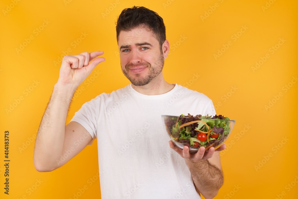 Upset young handsome Caucasian man holding a salad bowl against yellow wall shapes little gesture with hand demonstrates something very tiny small size. Not very much Upset young handsome Caucasian man holding a salad bowl against yellow wall shapes little gesture with hand demonstrates something very tiny small size. Not very much