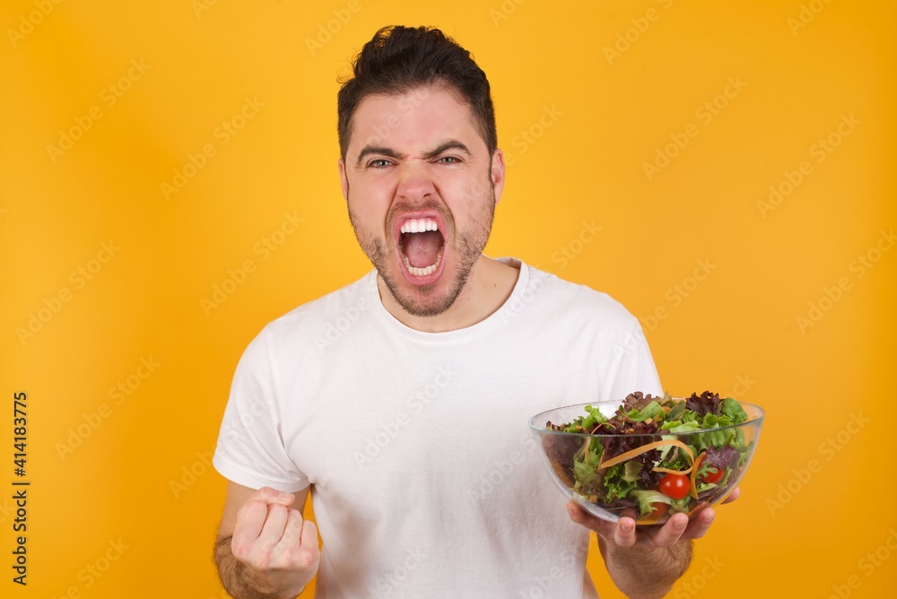 Joyful excited lucky young handsome Caucasian man holding a salad bowl against yellow wall cheering, celebrating success, screaming yes with clenched fists