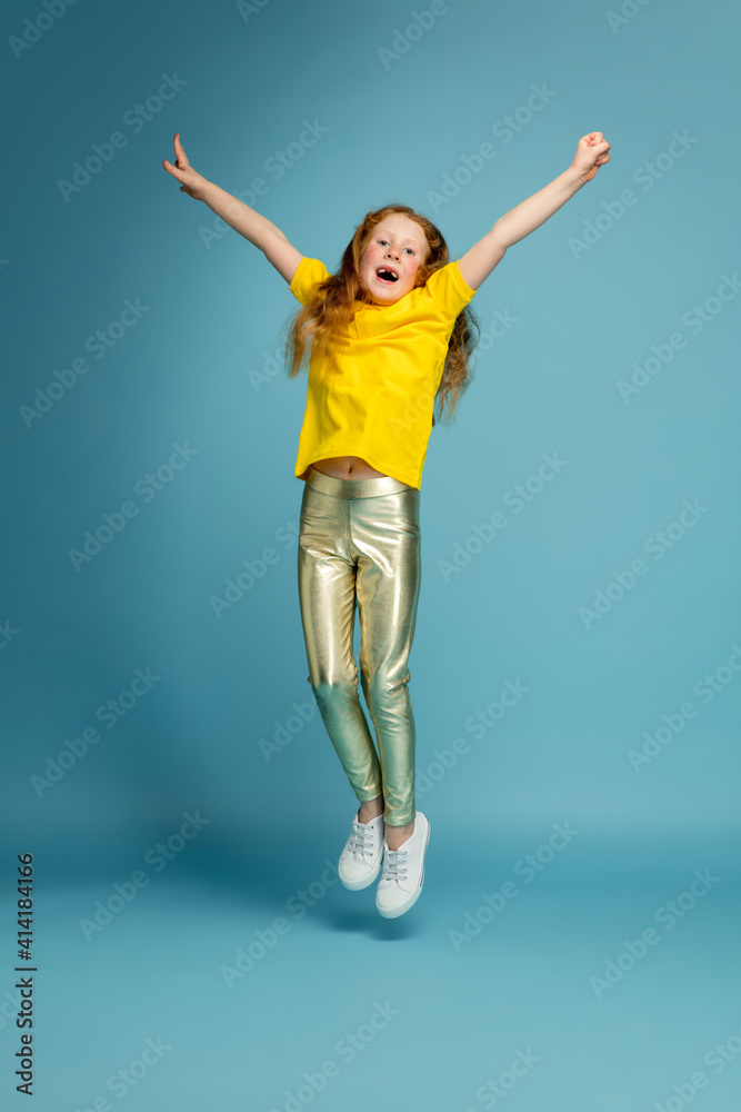 Jumping high. Happy, smiley redhair girl isolated on blue studio background with copyspace for ad. Looks happy, cheerful. Childhood, education, human emotions, facial expression concept. Jumping high. Happy, smiley redhair girl isolated on blue studio background with copyspace for ad. Looks happy, cheerful. Childhood, education, human emotions, facial expression concept.