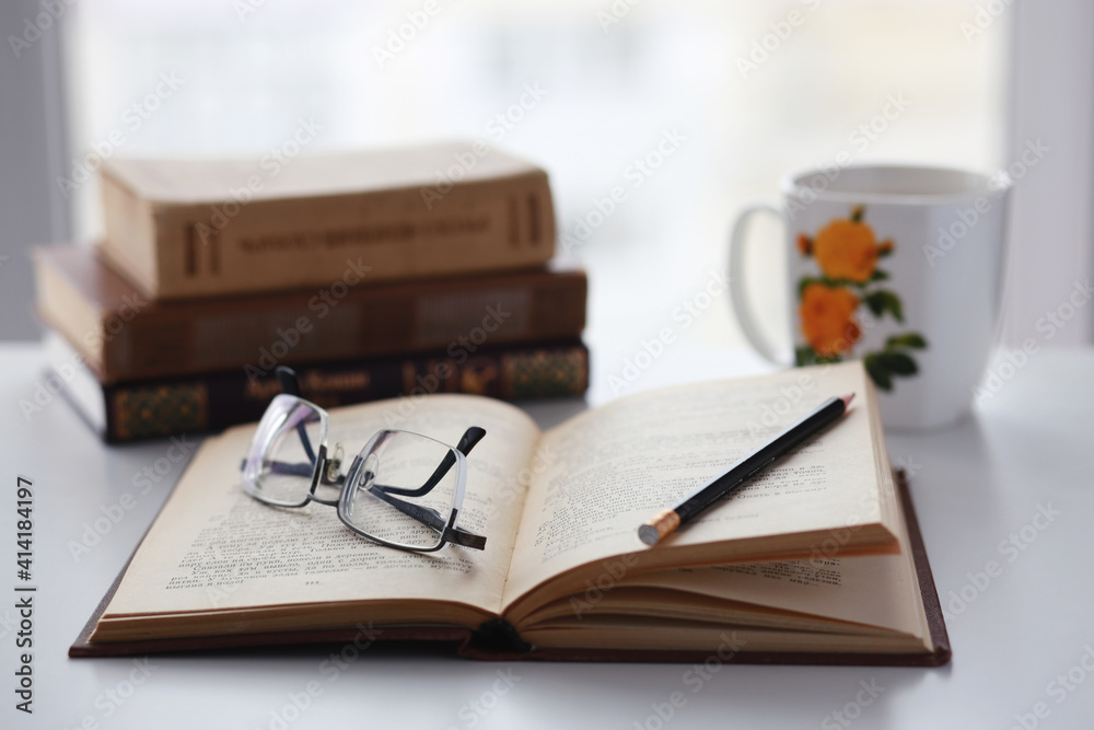 Naklejka premium Old books, glasses, pencil and cup of coffee on white table background against window light with copy space