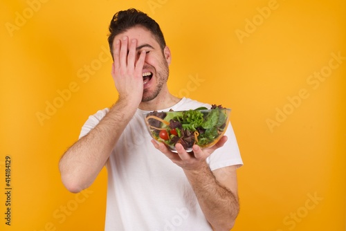 Charismatic carefree joyful young handsome Caucasian man holding a salad bowl against yellow wall likes laugh out loud not hiding emotions giggling hear funny hilarious joke chuckling facepalm.