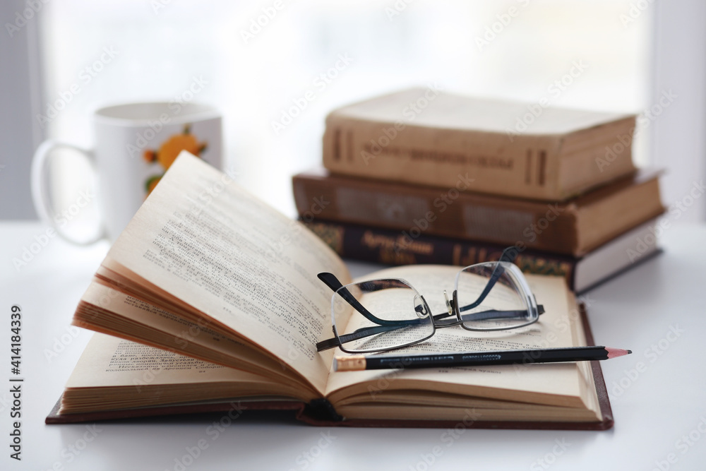 Old books, glasses, pencil and cup of coffee on white table background against window light with copy space. Vintage cozy warm mockup Old books, glasses, pencil and cup of coffee on white table background against window light with copy space. Vintage cozy warm mockup