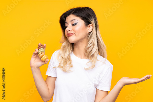 Teenager girl isolated on yellow background holding colorful French macarons and making doubts gesture