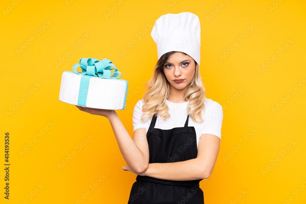 Teenager girl pastry holding a big cake isolated on yellow background keeping arms crossed Teenager girl pastry holding a big cake isolated on yellow background keeping arms crossed