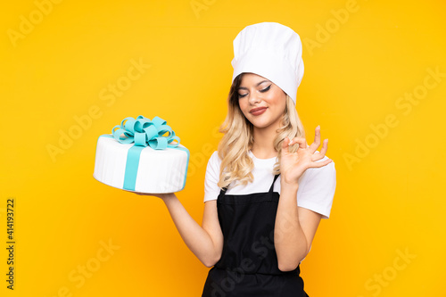 Teenager girl pastry holding a big cake isolated on yellow background showing an ok sign with fingers