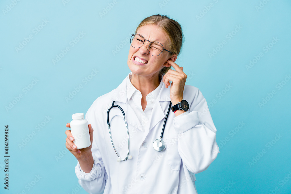 Young russian doctor woman holding pills bottle on blue covering ears with hands. Young russian doctor woman holding pills bottle on blue covering ears with hands.