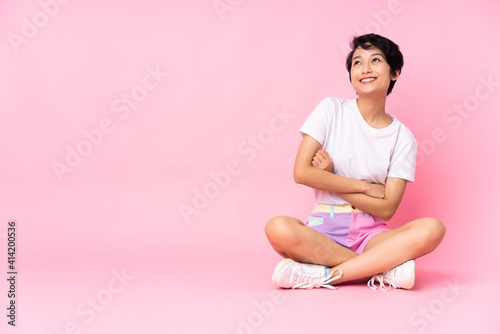 Young Vietnamese woman with short hair sitting on the floor over isolated pink background looking up while smiling