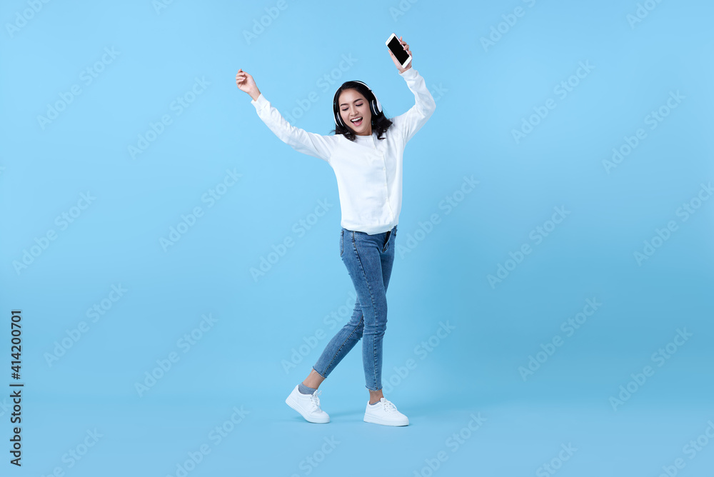Happy smiling Asian woman wearing wireless headphones listening to music and dancing on blue background.