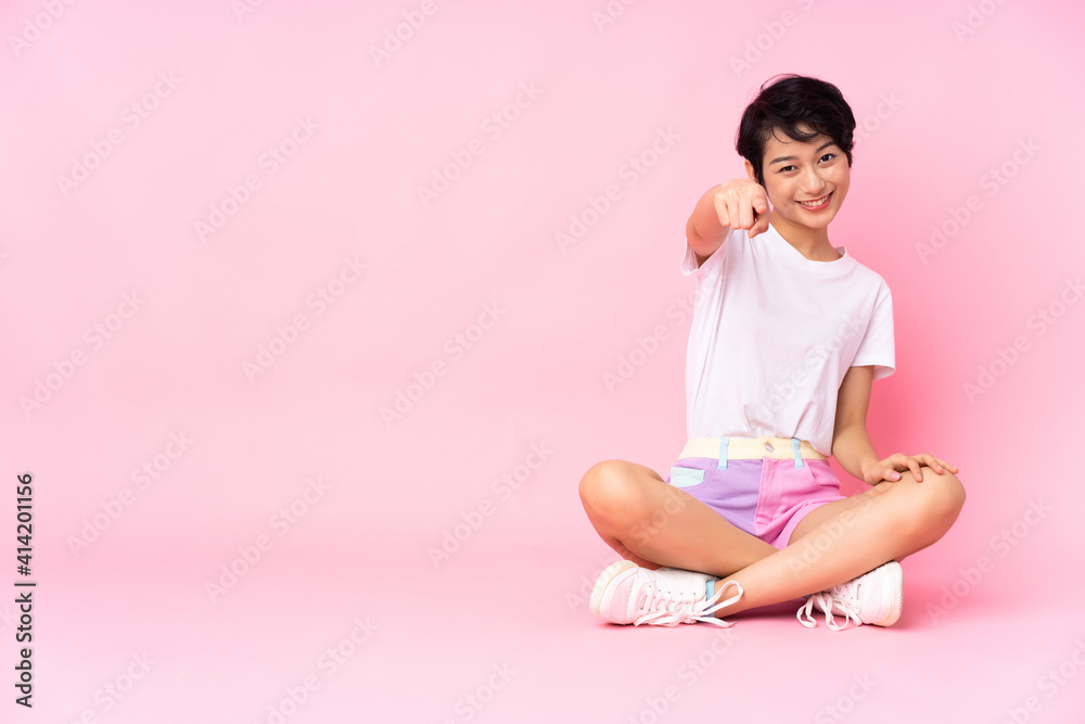 Young Vietnamese woman with short hair sitting on the floor over isolated pink background points finger at you with a confident expression Young Vietnamese woman with short hair sitting on the floor over isolated pink background points finger at you with a confident expression