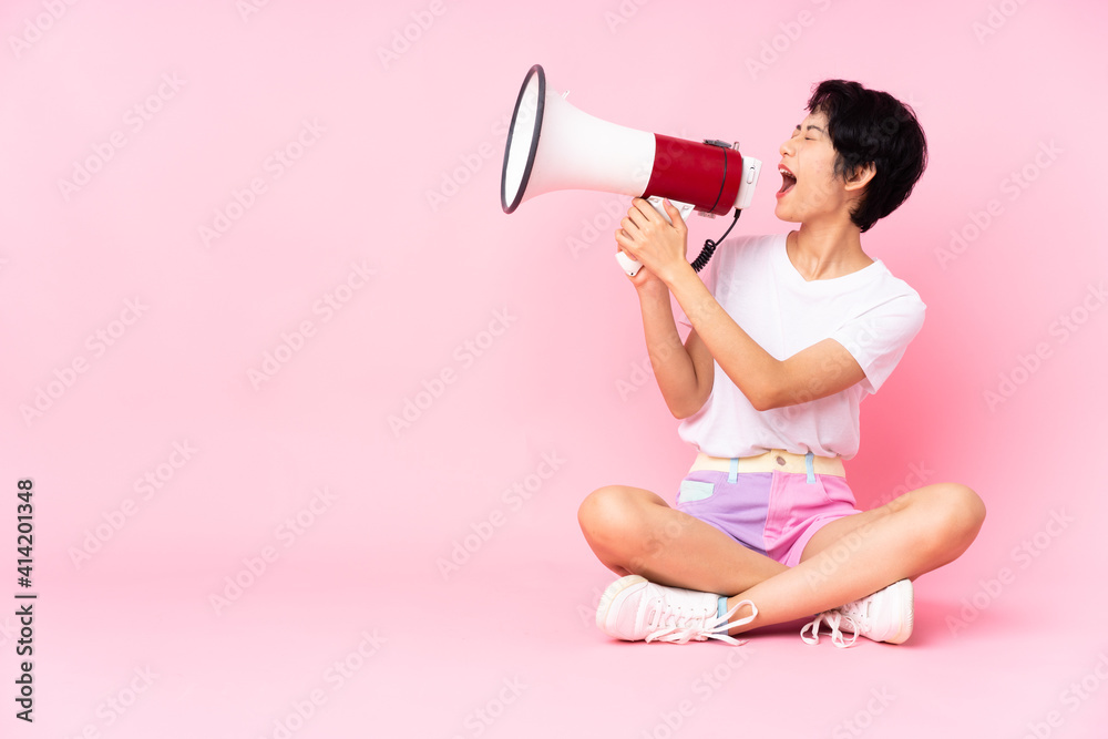 Young Vietnamese woman with short hair sitting on the floor over isolated pink background shouting through a megaphone Young Vietnamese woman with short hair sitting on the floor over isolated pink background shouting through a megaphone