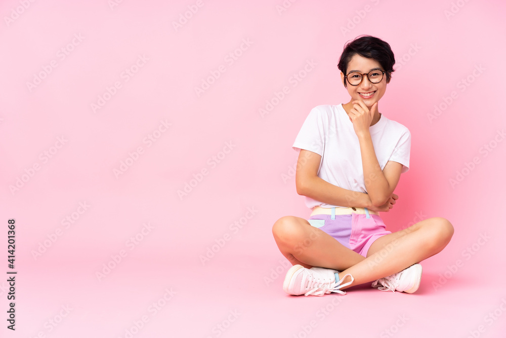 Young Vietnamese woman with short hair sitting on the floor over isolated pink background with glasses and smiling Young Vietnamese woman with short hair sitting on the floor over isolated pink background with glasses and smiling